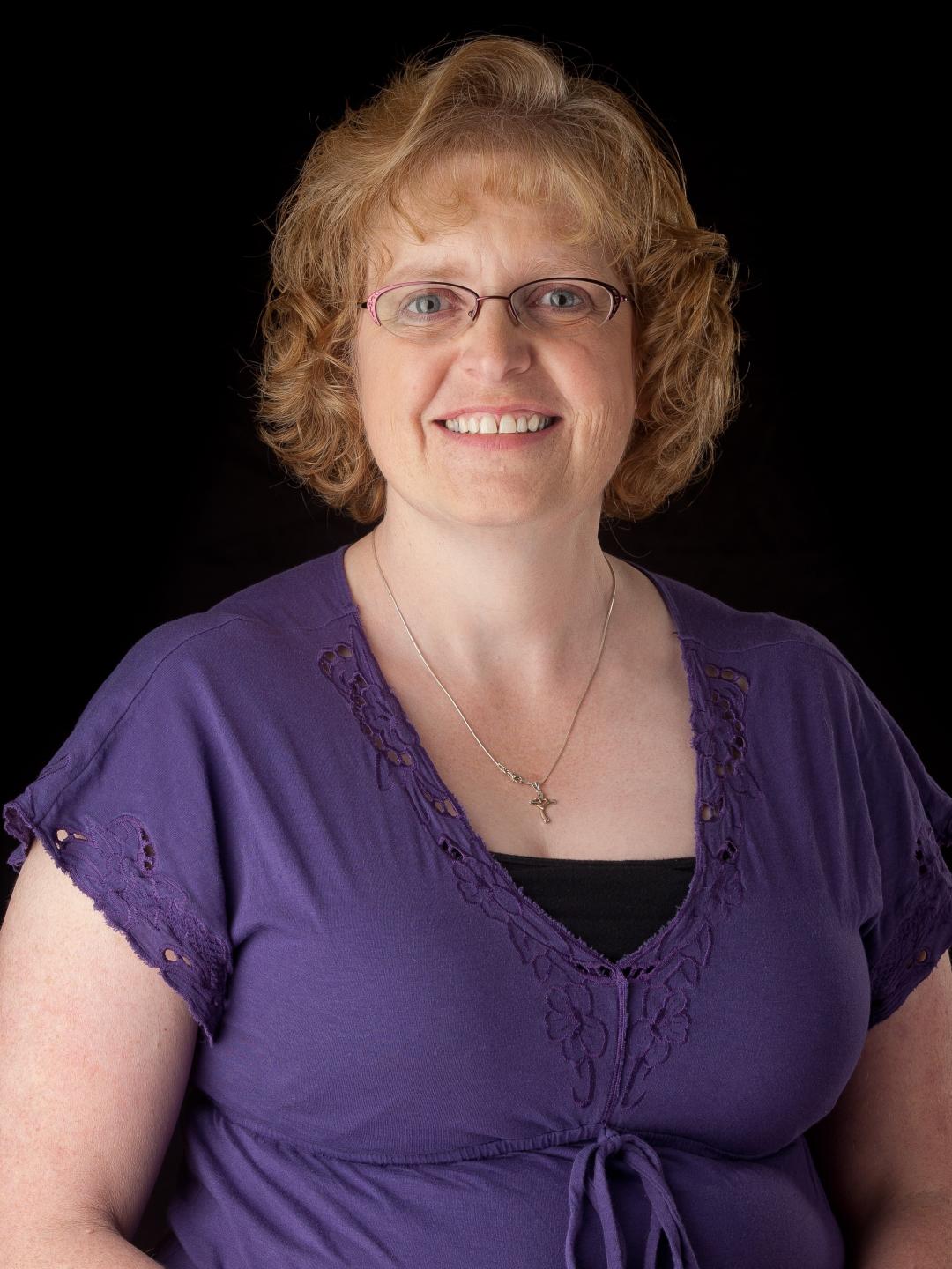 A woman with short red hair in front of a black background wearing a purple shirt.