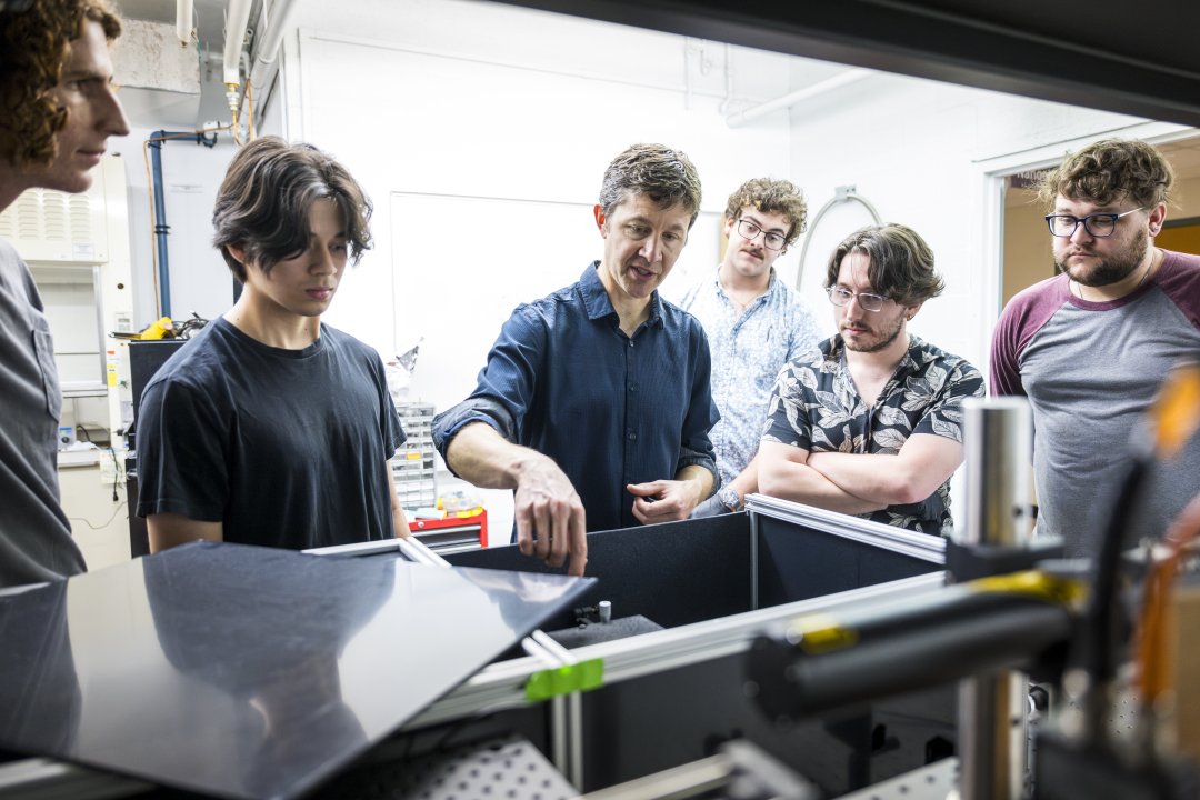 A man points to a piece of technology in a lab while a group of male students observe.