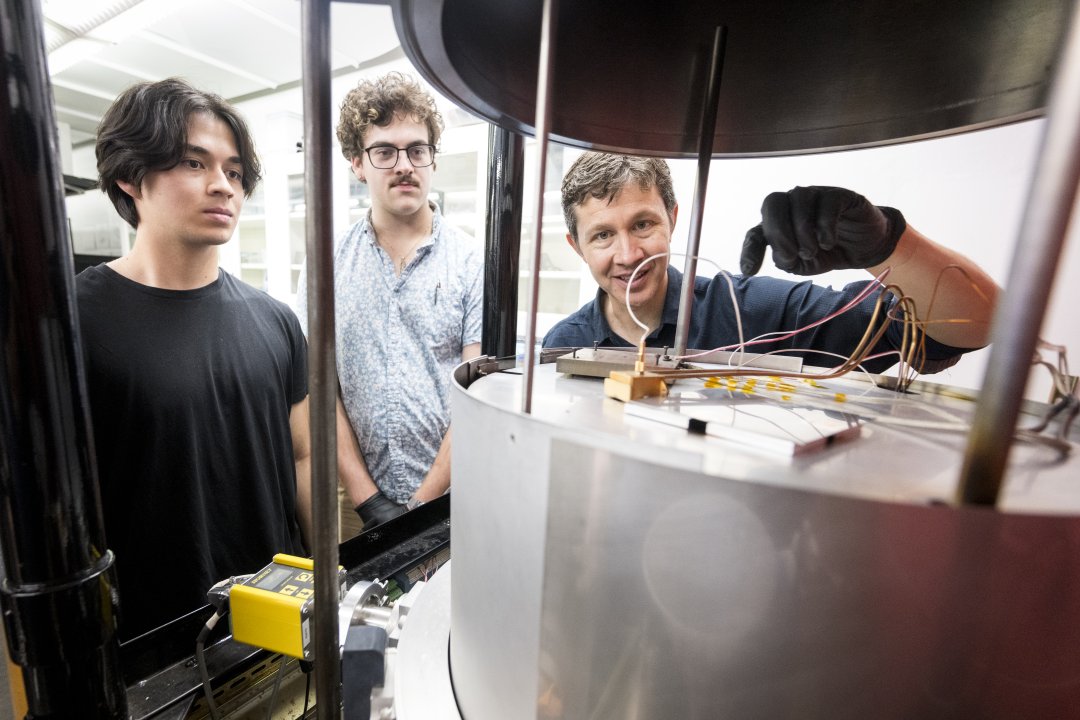 A man in a blue shirt with black gloves points at a machine while two male students observe.