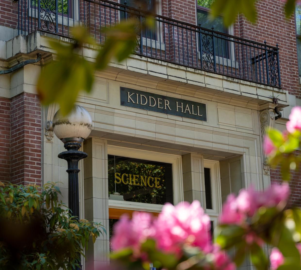 Photo of Kidder Hall from a low angle looking at a door with science written in the glass above the door, and Kidder Hall written in the stone above the glass. Blurred pink flowers appear in the foreground.