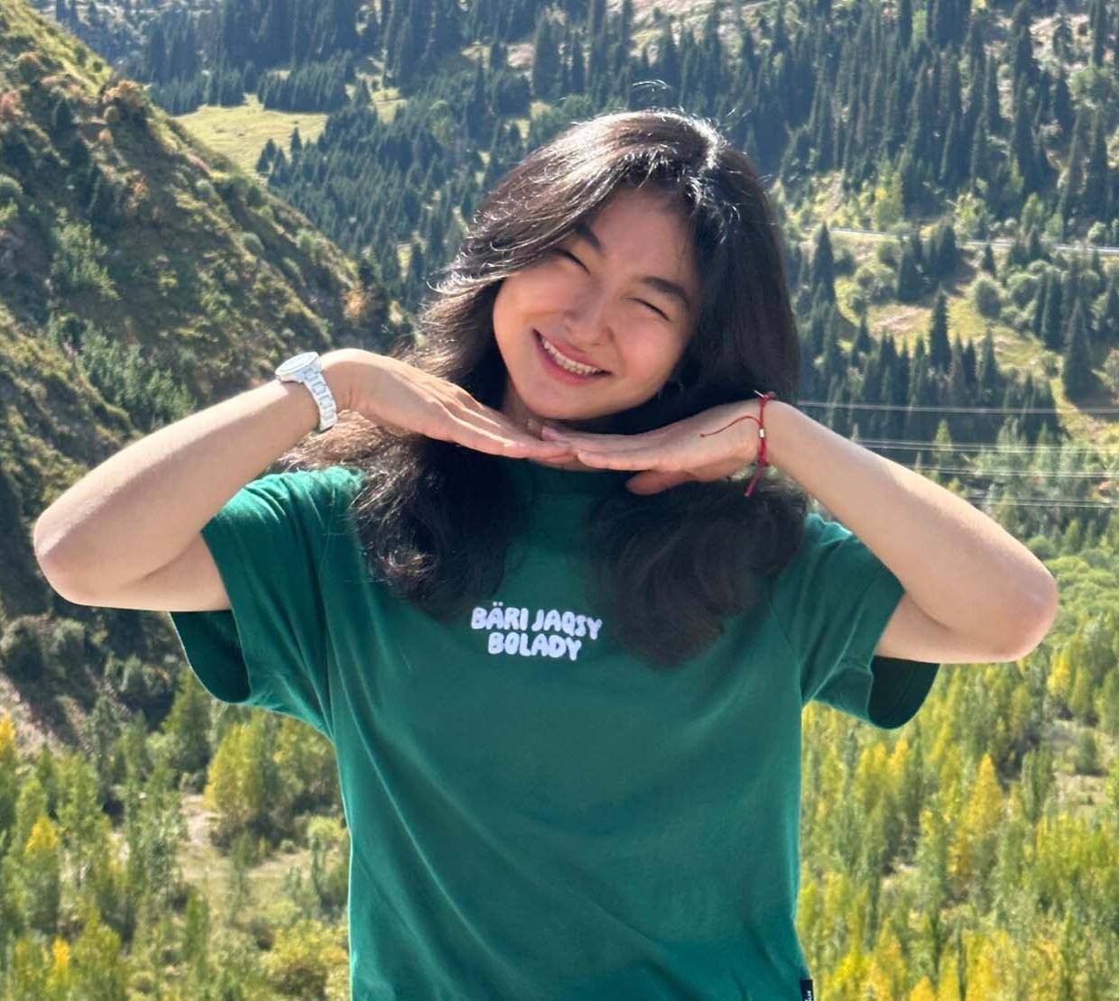 A woman with long black hair smiles, holding her hands around her face. Pine trees and shrubs blanket the mountains behind her.