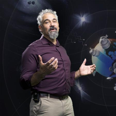 Astrophysicist Jeffrey Hazboun stands in front of an graphic of Earth surrounded by satellites and other cosmic bodies.