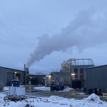 Under a cloudy sky, snow covers the ground of a small factory exterior as the building emits a long plume of smoke. 