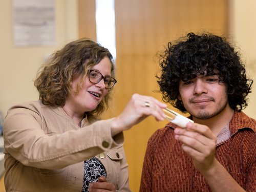 A student researcher looks at a vial of fruit flies with his smiling mentor