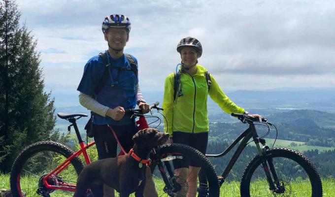 Courtney Rae Armour with husband and dog riding mountain bikes atop grassy hill