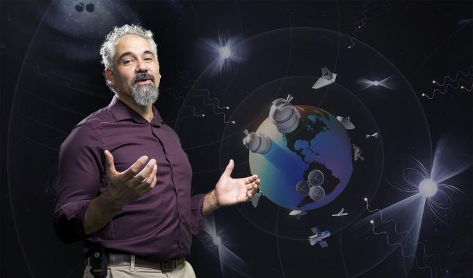 Astrophysicist Jeffrey Hazboun stands in front of an graphic of Earth surrounded by satellites and other cosmic bodies.