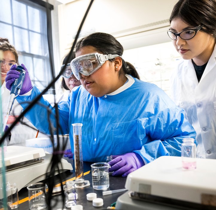 Students and staff clad in lab coats and glasses surround a lab table conducting experiments with pipettes, test tubes, beakers and other chemistry instruments in a lab at Oregon State University.